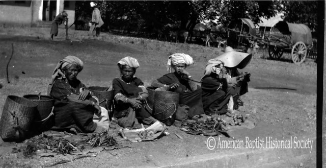 Four Pa-Oh women and one man (under the hat) in the 5-day bazaar at Taunggyi, Southern Shan State. Note the vegetables for sale, on dirty mats on the ground. See, also, the bracelets and heavy earrings, for these people carry all of their wealth with them. March 11, 1922. 