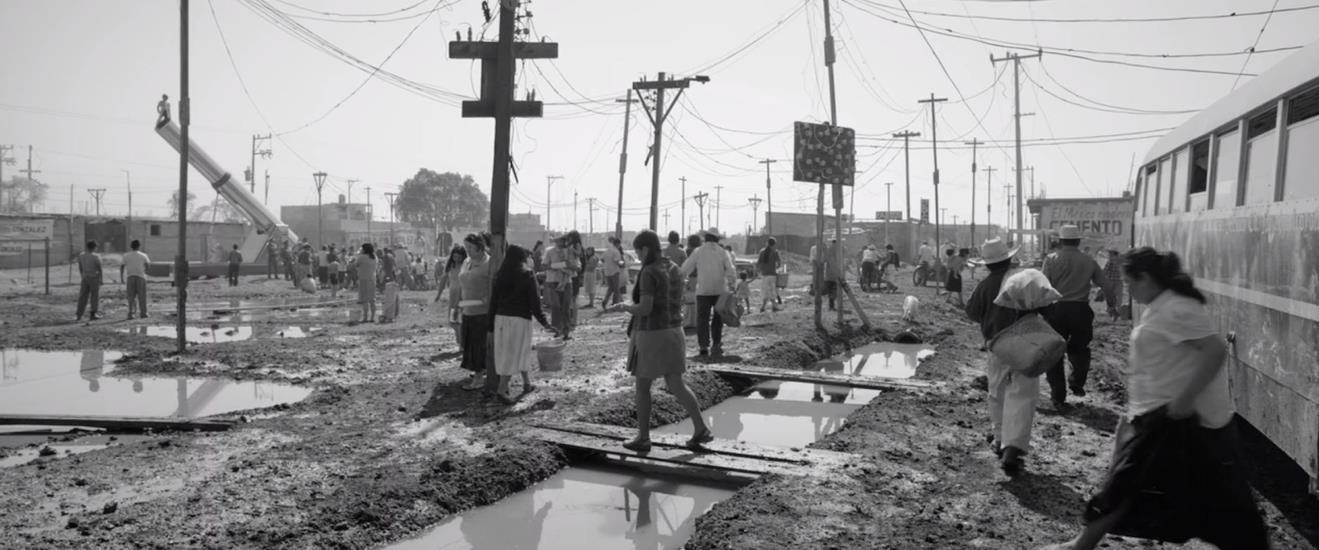 Figure 7: Cleo (Yalitza Aparicio) seeks Fermin (Jorge Antonio Guerrero) in the squalid Loma Colorada to inform him that she is having his baby. (Note the “human cannonball” in the background.) Figure 7: Cleo (Yalitza Aparicio) seeks Fermin (Jorge Antonio Guerrero) in the squalid Loma Colorada to inform him that she is having his baby. (Note the “human cannonball” in the background.)