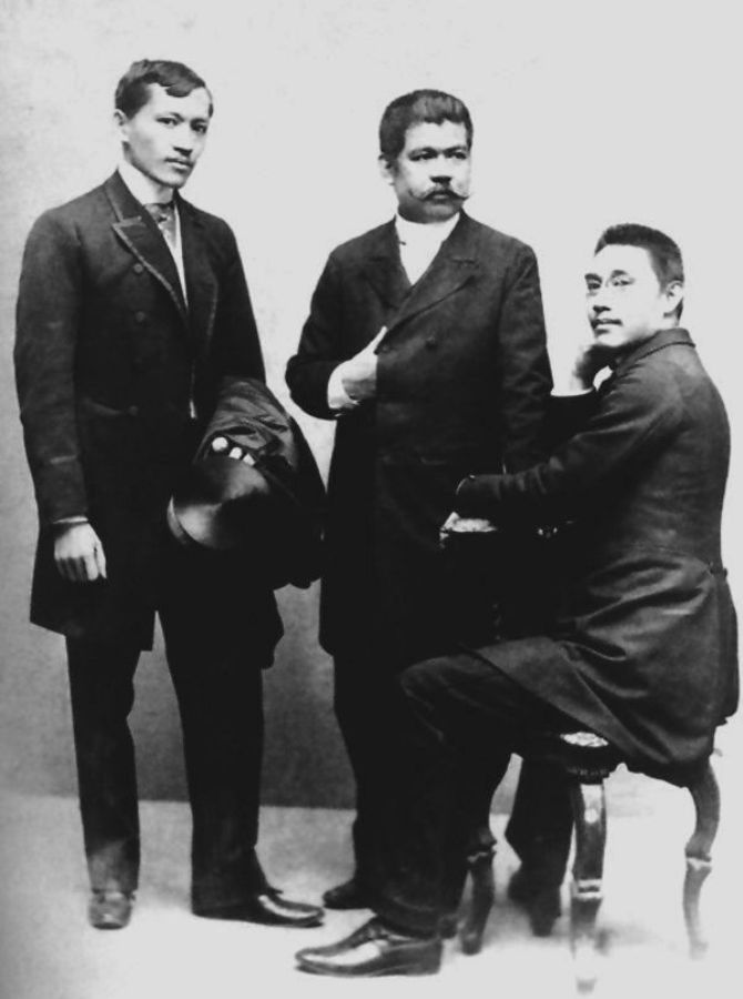Alt-text: “Three men in dark suits, one of whom is sitting on a chair.” Alt-text: “Three men in dark suits, one of whom is sitting on a chair.”