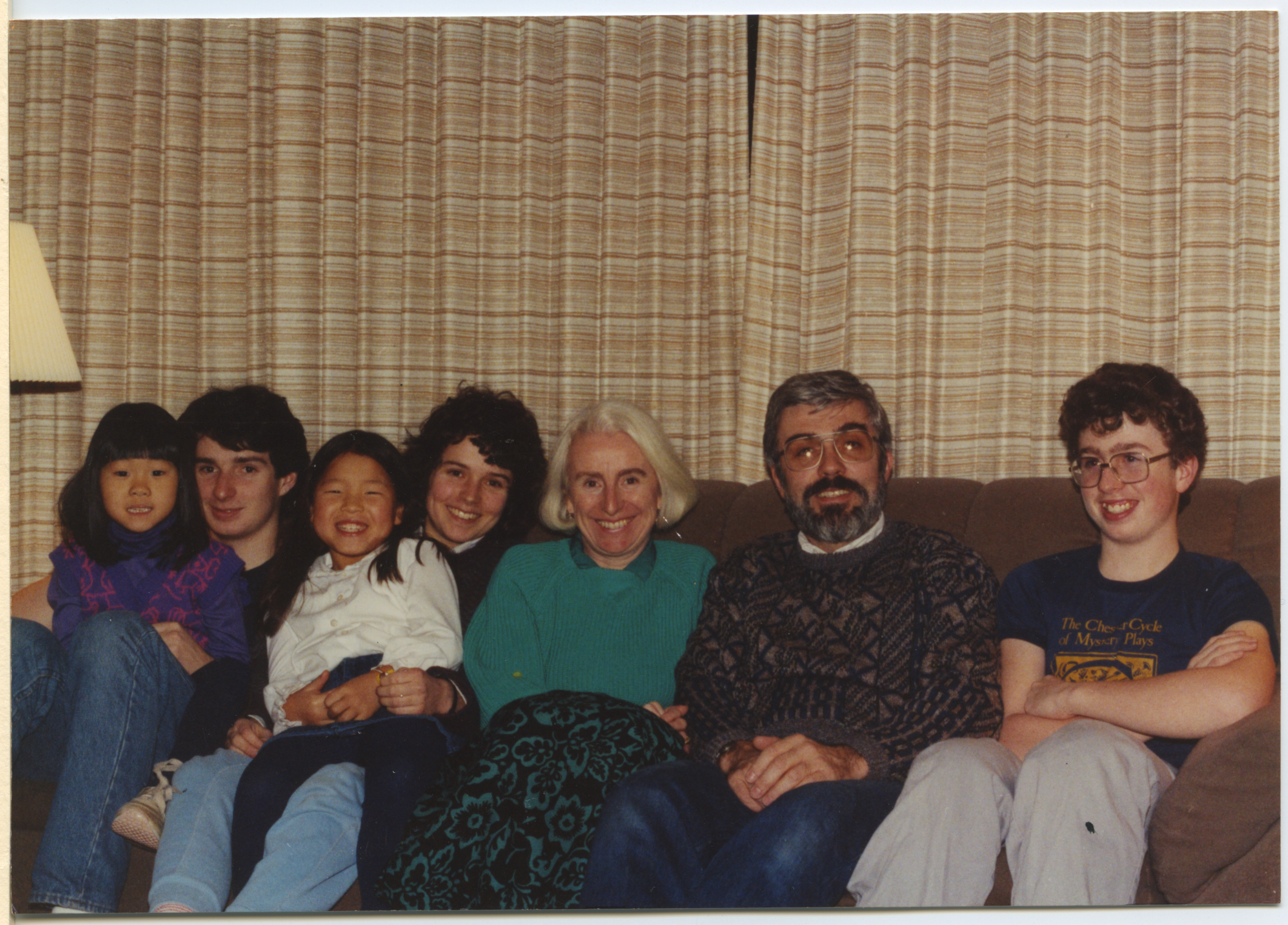 Alt-text: Five white people sit side by side on a couch smiling. Two Asian girls, also smiling, sit on the laps of two of them. Alt-text: Five white people sit side by side on a couch smiling. Two Asian girls, also smiling, sit on the laps of two of them.