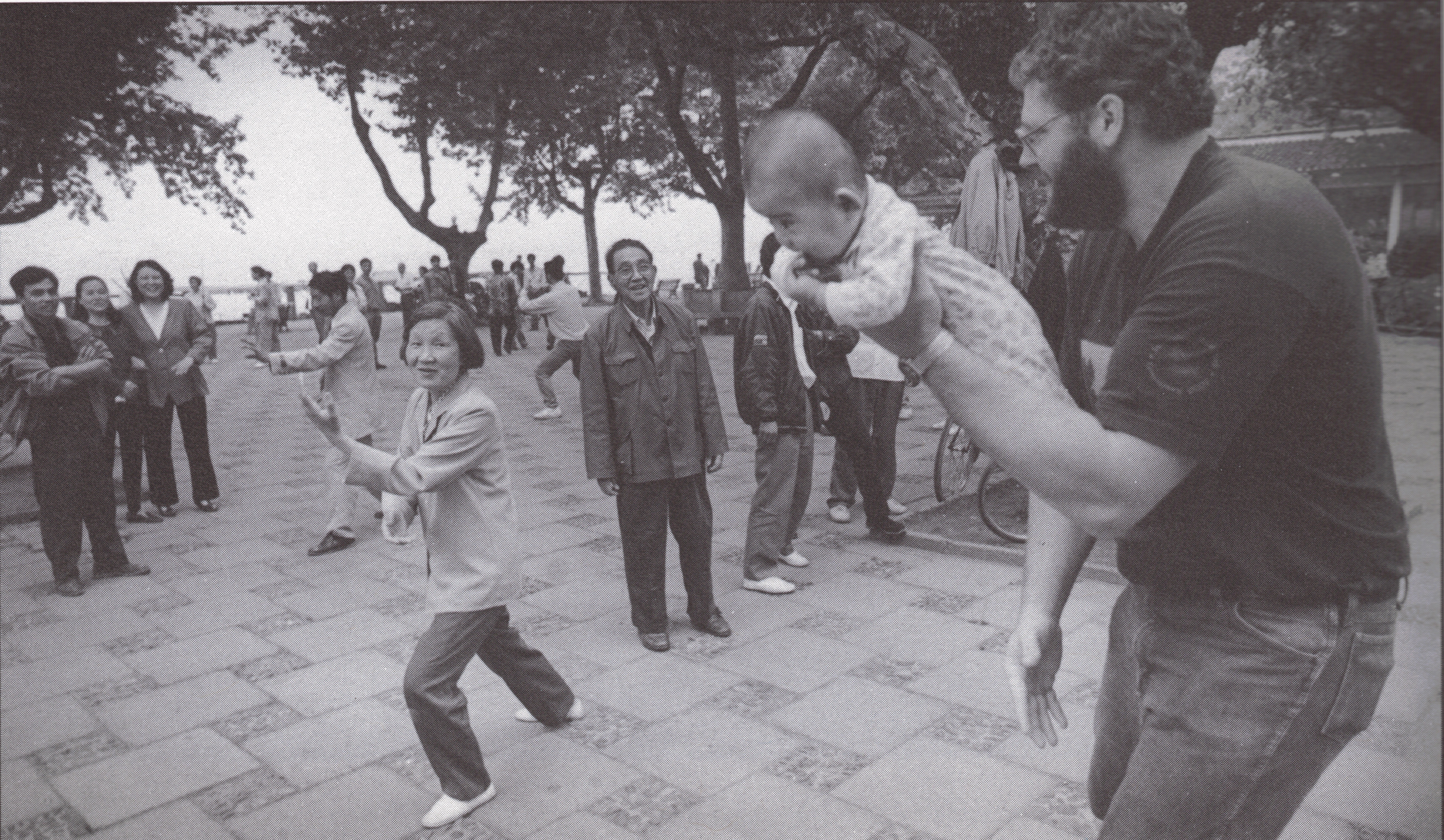 Alt-text: A black and white photograph of a Chinese woman doing tai chi in a park and a bearded white man holding a Chinese baby in his left arm, mimicking her pose. Chinese people stand around them looking amused. Alt-text: A black and white photograph of a Chinese woman doing tai chi in a park and a bearded white man holding a Chinese baby in his left arm, mimicking her pose. Chinese people stand around them looking amused.