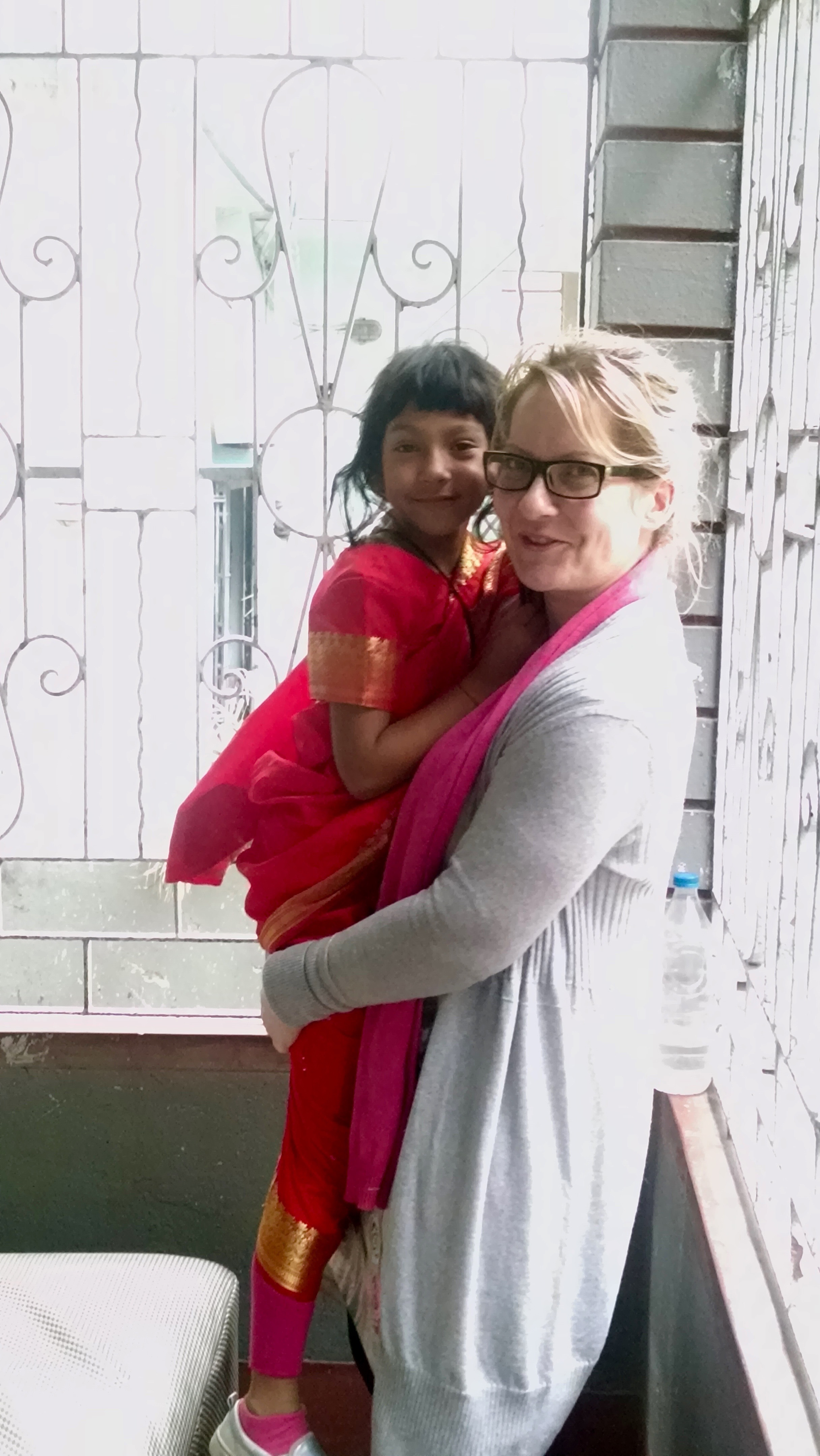 Alt-text: A white woman stands in a wrought-iron doorway smiling, holding a South Asian child. Alt-text: A white woman stands in a wrought-iron doorway smiling, holding a South Asian child.